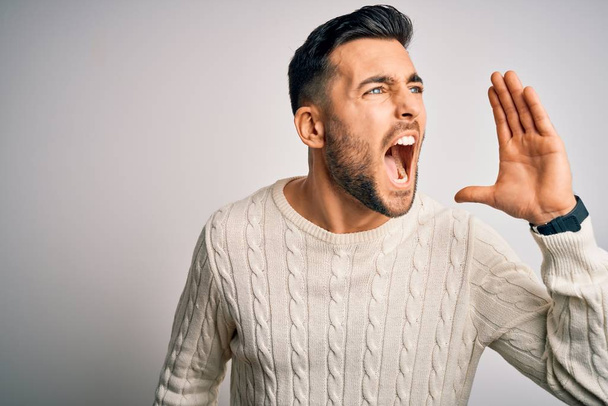 Young handsome man wearing casual sweater standing over isolated white background shouting and screaming loud to side with hand on mouth. Communication concept. - Zdjęcie, obraz