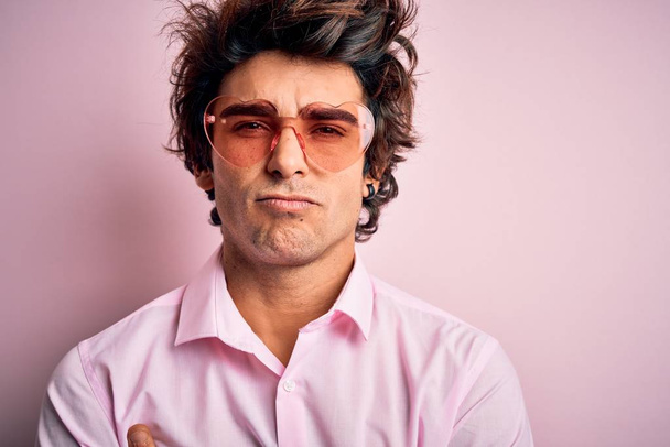 Young handsome man wearing glasses with heart form over isolated pink background skeptic and nervous, disapproving expression on face with crossed arms. Negative person. - Photo, Image