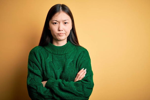 Young beautiful asian woman wearing green winter sweater over yellow isolated background skeptic and nervous, disapproving expression on face with crossed arms. Negative person. - Fotoğraf, Görsel