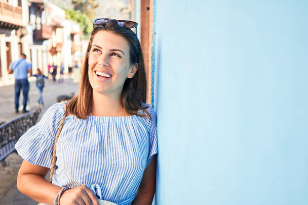 Красивая девушка опирается на голубую стену, молодая дружелюбная женщина улыбается счастливо в солнечный день лета
 - Фото, изображение