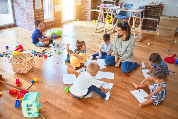 幼稚園のおもちゃの周りに紙や鉛筆を使って床に座っている子供たちの美しい先生とグループ - 写真・画像