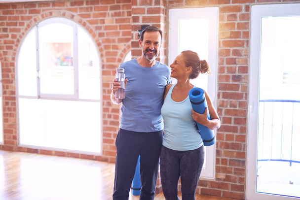 Schönes sportliches Paar mittleren Alters, das glücklich lächelt. mit Lächeln im Gesicht stehen und Matte im Fitnessstudio halten - Foto, Bild