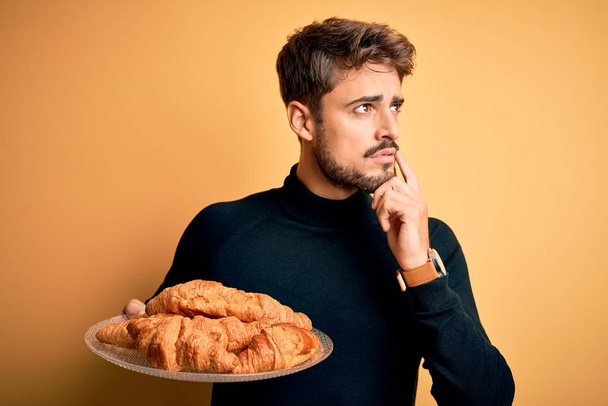 Giovane uomo con la barba piatto di tenuta con croissant in piedi sopra isolato sfondo giallo volto serio pensando alla domanda, idea molto confusa
 - Foto, immagini