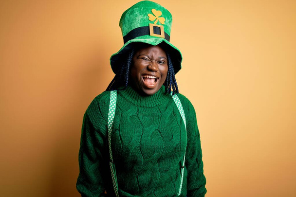 Африканская женщина plus size с брейдами в зеленой шляпе с клевером в день святого Патрика подмигивает, глядя в камеру с сексуальным выражением лица, веселым и счастливым лицом
. - Фото, изображение