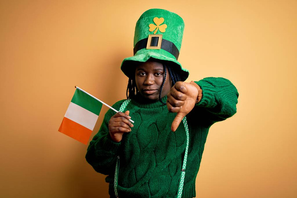 Plus la taille femme afro-américaine portant chapeau vert tenant drapeau irlandais sur saint patricks jour avec le visage en colère, signe négatif montrant aversion avec les pouces vers le bas, concept de rejet
 - Photo, image