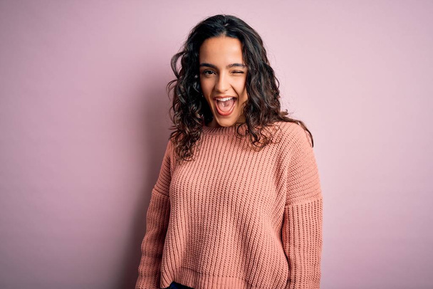 Young beautiful woman with curly hair wearing casual sweater over isolated pink background winking looking at the camera with sexy expression, cheerful and happy face. - Foto, Bild