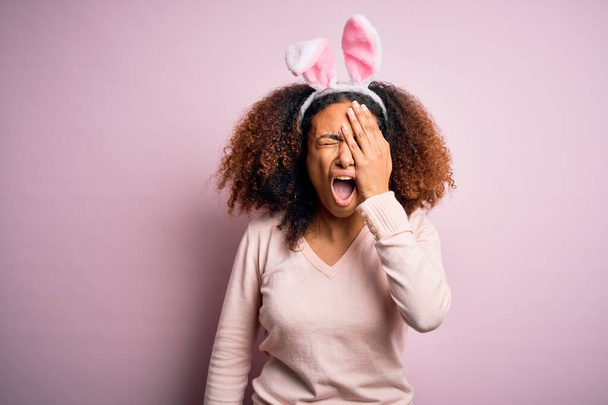 Jeune femme afro-américaine aux cheveux afro portant des oreilles de lapin sur fond rose bâillement fatigué couvrant la moitié du visage, des yeux et de la bouche avec la main. Le visage souffre dans la douleur
. - Photo, image