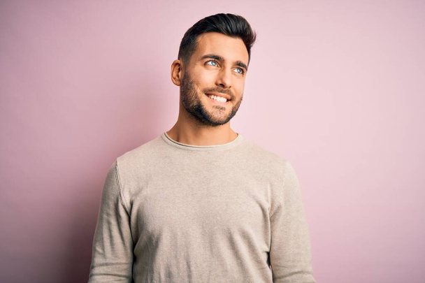 Young handsome man wearing casual sweater standing over isolated pink background looking away to side with smile on face, natural expression. Laughing confident. - Foto, afbeelding