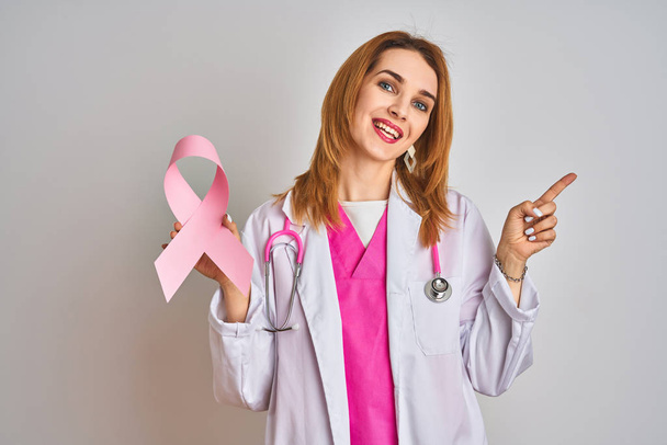 Pelirroja mujer médica caucásica sosteniendo cinta de cáncer rosa sobre fondo aislado muy feliz señalando con la mano y el dedo hacia el lado
 - Foto, Imagen