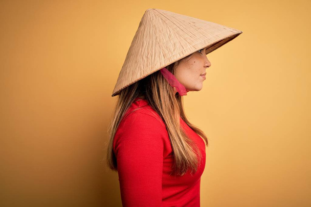 Młoda blondynka w tradycyjnym azjatyckim ryżu paddy słomy kapelusz na żółtym tle patrząc na bok, relaks profil pozować z naturalną twarz z pewnym uśmiechem. - Zdjęcie, obraz
