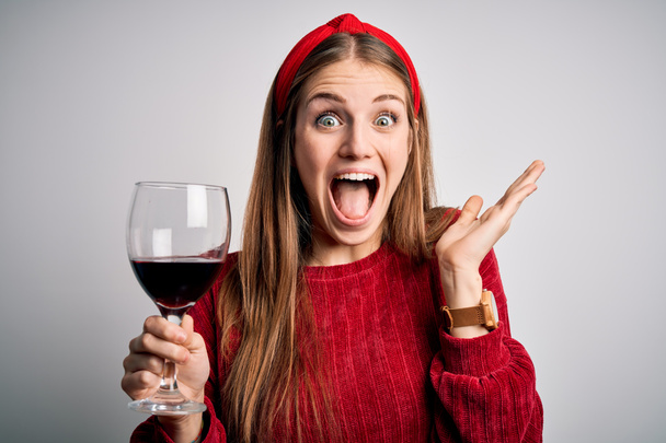 Genç, güzel, kızıl saçlı bir kadın izole edilmiş beyaz arka planda bir bardak kırmızı şarap içiyor. Mutlu ve heyecanlı. Kazanan ifade zaferi kutluyor. - Fotoğraf, Görsel