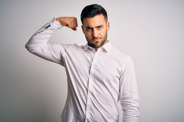 Νέος όμορφος άνδρας φορώντας κομψό πουκάμισο στέκεται πάνω από απομονωμένο λευκό φόντο Ισχυρός άνθρωπος δείχνει μπράτσο μυών, αυτοπεποίθηση και περήφανος για την εξουσία - Φωτογραφία, εικόνα