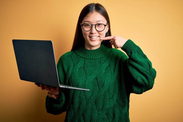 Молода азіатська бізнес-леді в окулярах і працює з використанням комп'ютерного ноутбука усміхнений веселий показ і вказівка пальцями зубів і рота. Концепція стоматологічного здоров'я
. - Фото, зображення