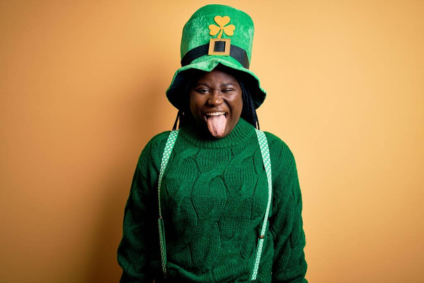 Plus Afrikaans-Amerikaanse vrouw met vlechten met groene hoed met klaver op St Patrick 's Day, die zijn tong uitsteekt, blij met grappige uitdrukking. Emotieconcept. - Foto, afbeelding