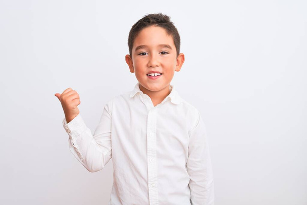 Mooie jongen draagt elegante shirt staan over geïsoleerde witte achtergrond glimlachen met gelukkig gezicht kijken en wijzen naar de zijkant met duim omhoog. - Foto, afbeelding