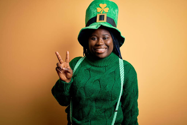 Más tamaño mujer afroamericana con trenzas con sombrero verde con trébol en st patricks día mostrando y señalando hacia arriba con los dedos número dos mientras sonríe confiado y feliz
. - Foto, imagen