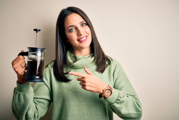 Молодая женщина с голубыми глазами делает кафе с помощью кофеварки стоя на белом фоне очень счастливо указывая рукой и пальцем
 - Фото, изображение