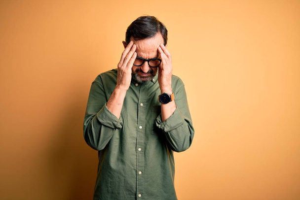 Middle age hoary man wearing casual green shirt and glasses over isolated yellow background with hand on head for pain in head because stress. Suffering migraine. - Foto, immagini