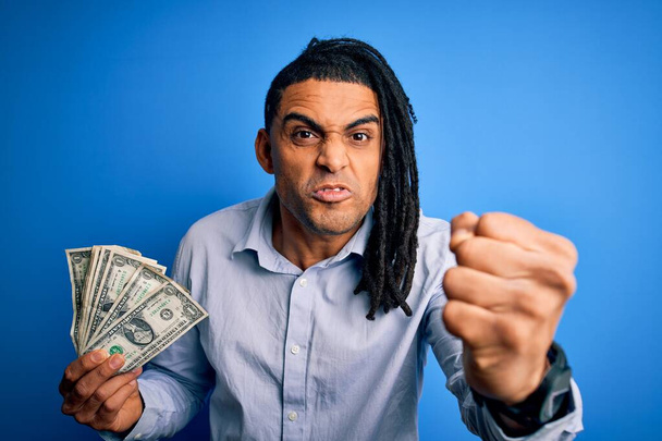 jovem afro americano africano homem com dreadlocks segurando dólares sobre fundo azul irritado e frustrado gritando com raiva, louco e gritando com a mão levantada, conceito de raiva
 - Foto, Imagem