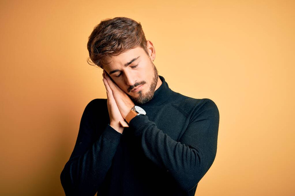 Young handsome man with beard wearing turtleneck sweater standing over yellow background sleeping tired dreaming and posing with hands together while smiling with closed eyes. - Photo, Image