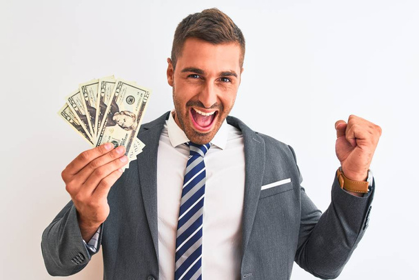 Young handsome business man holding bunch of dollars banknotes over isolated background screaming proud and celebrating victory and success very excited, cheering emotion - Photo, Image