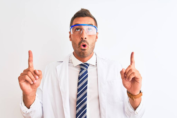 Jonge knappe wetenschapper draagt een veiligheidsbril over geïsoleerde achtergrond verbaasd en verrast omhoog kijkend en wijzend met vingers en opgeheven armen. - Foto, afbeelding