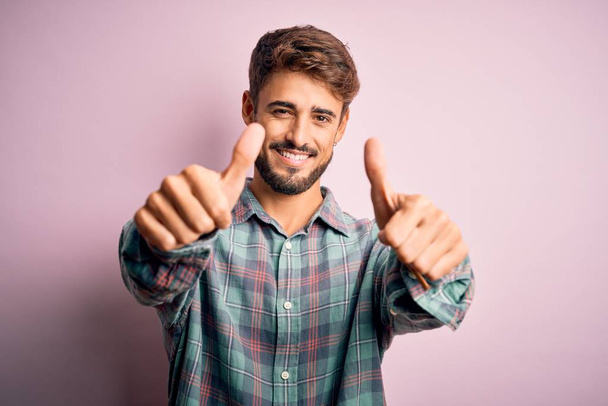 Young handsome man with beard wearing casual shirt standing over pink background approving doing positive gesture with hand, thumbs up smiling and happy for success. Winner gesture. - Foto, Imagen