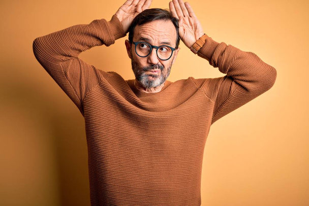 Middle age hoary man wearing brown sweater and glasses over isolated yellow background Doing bunny ears gesture with hands palms looking cynical and skeptical. Easter rabbit concept. - Photo, Image
