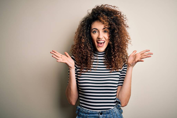 Young beautiful woman with curly hair and piercing wearing casual striped t-shirt celebrating crazy and amazed for success with arms raised and open eyes screaming excited. Winner concept - Foto, imagen