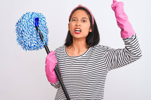 Young chinese cleaner woman wearing gloves holding mop over isolated white background annoyed and frustrated shouting with anger, crazy and yelling with raised hand, anger concept - Foto, Imagem