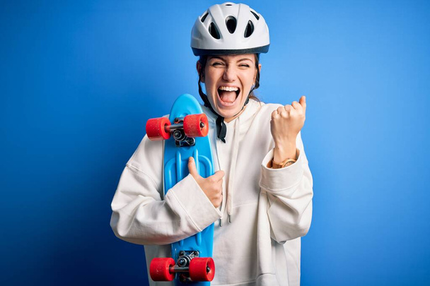 Jonge mooie roodharige skateboarder vrouw draagt veiligheidshelm houden skate schreeuwen trots en vieren overwinning en succes zeer opgewonden, juichen emotie - Foto, afbeelding