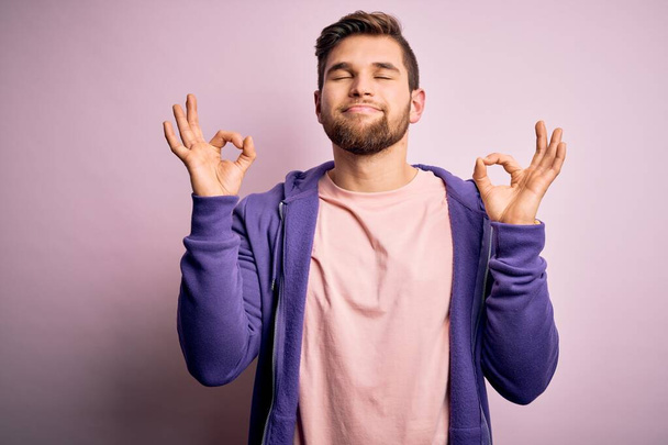 Young blond man with beard and blue eyes wearing purple sweatshirt over pink background relax and smiling with eyes closed doing meditation gesture with fingers. Yoga concept. - Fotoğraf, Görsel