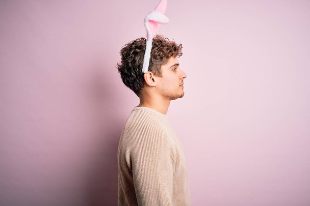 Joven hombre guapo rubio con orejas de conejo de Pascua sobre fondo rosa aislado mirando a un lado, pose de perfil relajado con cara natural con sonrisa confiada
. - Foto, Imagen