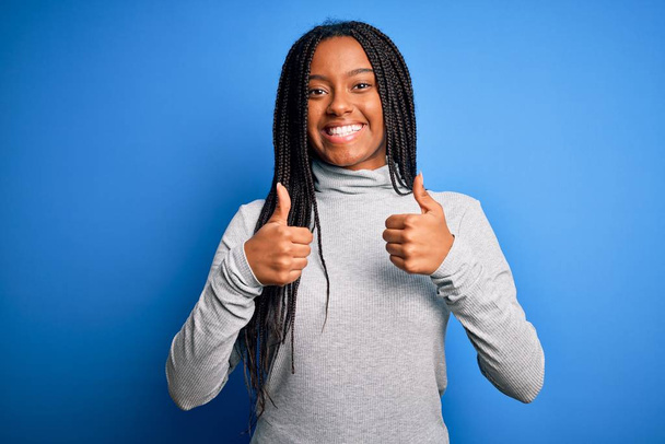 青い隔離された背景の成功の印の上のカジュアルなタートルネックを身に着けている若いアフリカ系アメリカ人の女性は手で肯定的なジェスチャーをする、親指を笑顔と幸せ。陽気な表情と勝者のジェスチャー. - 写真・画像