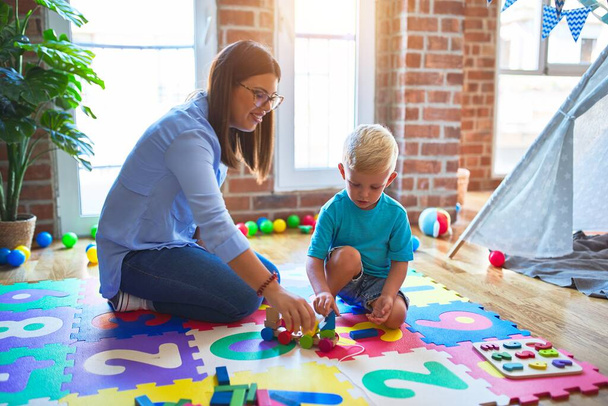 Young caucasian child playing at playschool with teacher. Mother and son playing with wooden pieces train at playroom - Photo, Image
