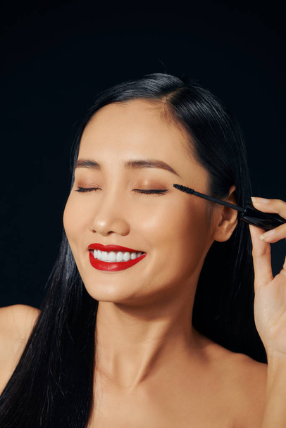 Κοντινό πλάνο πορτρέτο του ωραία όμορφη χαριτωμένο γλυκό ελκυστική χαρούμενη θετική ασιατική γυναίκα κρατώντας στα χέρια εφαρμόζοντας μοντέρνα μαύρη μάσκαρα απομονωμένη - Φωτογραφία, εικόνα