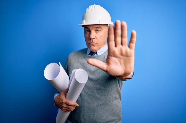 Middle age handsome grey-haired architect man wearing safety helmet holding blueprints doing stop sing with palm of the hand. Warning expression with negative and serious gesture on the face. - Photo, Image