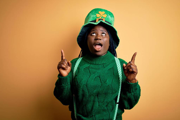 Plus size mulher americana africana com tranças usando chapéu verde com trevo no dia st patricks espantado e surpreso olhando para cima e apontando com os dedos e braços levantados
. - Foto, Imagem
