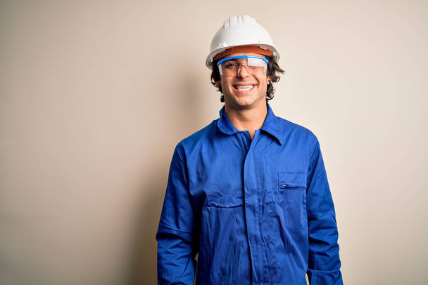 Young constructor man wearing uniform and security helmet over isolated white background winking looking at the camera with sexy expression, cheerful and happy face. - Photo, Image