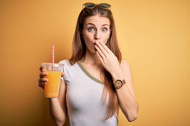 Young beautiful redhead woman drinking healthy orange juice over yellow background cover mouth with hand shocked with shame for mistake, expression of fear, scared in silence, secret concept - Photo, Image