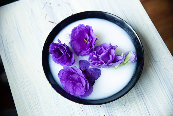 hermosas flores artificiales de eustoma púrpura en un plato negro con leche en un fondo de madera. - Foto, imagen