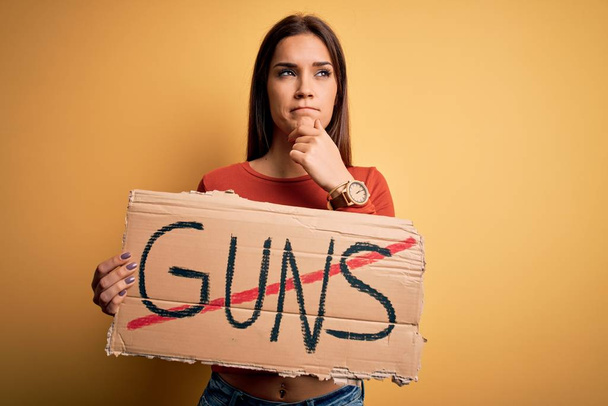 Νεαρή όμορφη ακτιβίστρια γυναίκα ζητά την ειρήνη κρατώντας πανό με όπλα stop μήνυμα σοβαρό πρόσωπο σκέφτεται την ερώτηση, πολύ μπερδεμένη ιδέα - Φωτογραφία, εικόνα