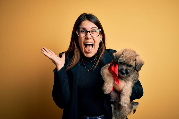 Young beautiful brunette woman holding cute puppy pet over isolated yellow background very happy and excited, winner expression celebrating victory screaming with big smile and raised hands - 写真・画像