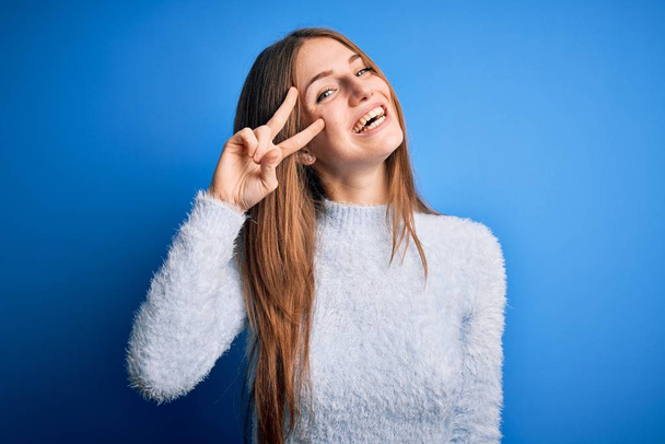 Young beautiful redhead woman wearing casual sweater over isolated blue background Doing peace symbol with fingers over face, smiling cheerful showing victory - Foto, Imagen
