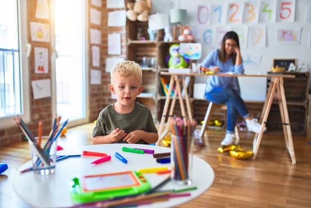 Nuori valkoihoinen lapsi leikkii leikkikoulussa opettajan kanssa. Äiti ja poika leikkihuoneessa piirustus piirtää väri kyniä, nuori nainen taustalla istuu pöydällä
. - Valokuva, kuva
