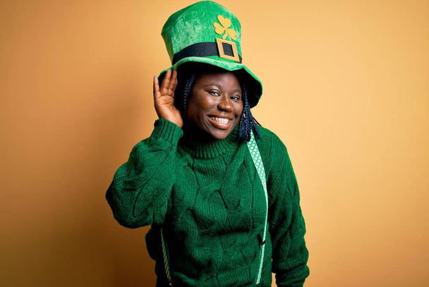 Plus size african american woman with braids wearing green hat with clover on st patricks day smiling with hand over ear listening an hearing to rumor or gossip. Deafness concept. - Photo, Image