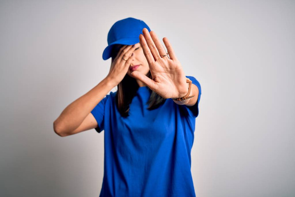 Young delivery woman with blue eyes wearing cap standing over blue background covering eyes with hands and doing stop gesture with sad and fear expression. Embarrassed and negative concept. - Foto, imagen