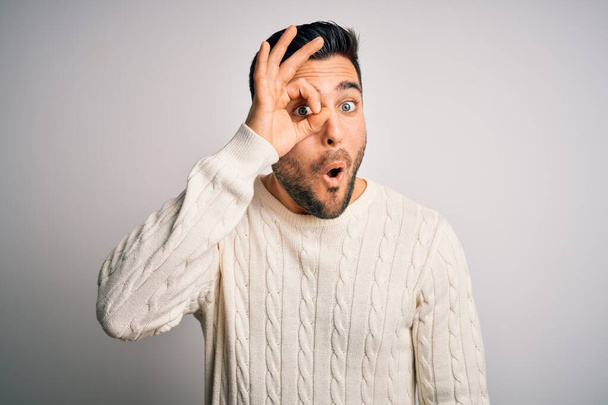 Young handsome man wearing casual sweater standing over isolated white background doing ok gesture shocked with surprised face, eye looking through fingers. Unbelieving expression. - Valokuva, kuva