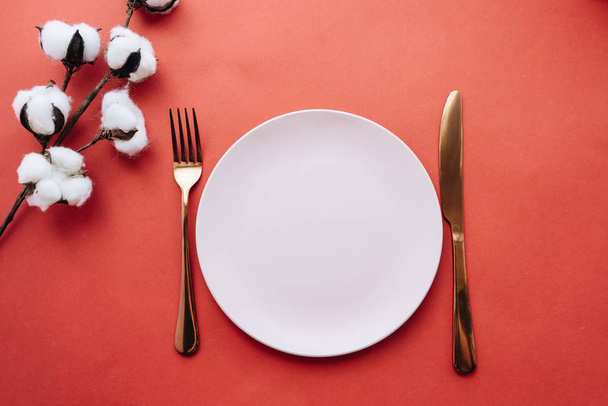 biały talerz, widelec, nóż i kwiaty bawełny na czerwonym stole, eleganckie zastawy stołowe  - Zdjęcie, obraz