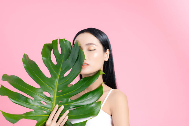 Young Asian woman holding a palm leaf to hide a half face on pink background. Beauty concept. - Фото, изображение